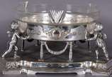 Centro de mesa de metal prateado na forma de bo... - Centro de mesa de metal prateado na forma de bowl com parte interna de cristal e laterais com guirlandas em relevo, sustentado por figuras de an...