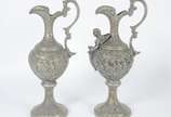 Par de ânforas de metal com anjos, flores e fol... - Par de ânforas de metal com anjos, flores e folhagens em relevo. Uma delas com aplique de figura alada estilizada. Desgastes no metal.
