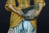 Estatueta de porcelana policromada representand... - Estatueta de porcelana policromada representando moça com cesto de uvas
