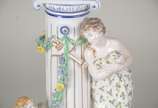 Fruteira de porcelana alemã policromada, base c... - Fruteira de porcelana alemã policromada, base com figura feminina recostada em coluna olhando para figura angelical com guirlandas florais, prat...