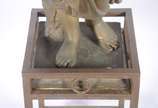 Estatueta de metal patinado representando figur... - Estatueta de metal patinado representando figura feminina com pássaro, acompanha coluna com tampo de vidro (metal com oxidação e desgastes).