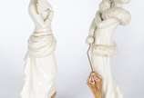 Par de estatuetas de porcelana portuguesa Época... - Par de estatuetas de porcelana portuguesa Época" nas cores branca e dourado, representando damas de época com chapéus, leque e sombrinha. Douraç...