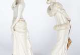 Par de estatuetas de porcelana portuguesa Época... - Par de estatuetas de porcelana portuguesa Época" nas cores branca e dourado, representando damas de época com chapéus, leque e sombrinha. Douraç...