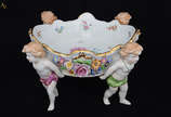Fruteira de porcelana alemã Schierholz , sendo ... - Fruteira de porcelana alemã Schierholz , sendo prato central sustentando por quatro figuras angelicais em policromia, uma delas com restauro, al...