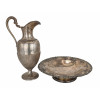 <p>Lote com 2 peças, composto por centro de mesa e jarra em metal prateado. Centro de mesa de formato circular, com borda vazada, ornamentação em relevo, sobre pedestal circular, medindo 29 cm de diâmetro e jarra de linhas clássicas, com corpo canelado na parte inferior, bico vertedor e alça sinuosa, medindo 30 cm de altura</p>