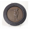 <p>Prato de bronze representando figura de Discóbolo, com assinatura não identificada, moldura de madeira entalhada. Diam. 28cm (total). Europa, início séc. XX</p>