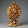 RELICÁRIO DE PAREDE<br />Brasil/Portugal, Século XIX — Estilo Barroco/Rococó<br />Placa em fibra de madeira e gesso estucado | Pintura manual e douração a folha de ouro sobre bolo armênio bordeaux<br />Medidas: 68 x 38 x 16 cm<br /><br />Imponente relicário parietal de devoção privada, executado no gosto barroco que dominou a arte sacra luso-brasileira do Oitocentos. Ricamente trabalhado à mão e douração a folha de ouro aplicada sobre bolo armênio bordeaux — técnica tradicional que confere calor e profundidade ao brilho metálico.<br /><br />Ao centro, óculo circular no formato de antigo hostiário, ladeado por parras e ramos de trigo em alto-relevo — símbolos eucarísticos do Vinho e do Pão, Corpo e Sangue de Cristo. A moldura raiada, em explosão de raios, remete à glória divina, e é encimada por cruz latina. Na base, cabeça de querubim alado em carnação policromada, guardião celeste do sacrário. É a teologia do Barroco materializada: drama, ouro e pedagogia visual para os fiéis.