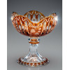 CRISTAL KARLOVY VARY — KARLSBAD<br />Boêmia, República Tcheca | Meados do séc. XX | Cristal doublé âmbar<br />Baleiro / Compoteira com pé | 17,1 x 16 x 16 cm | 932 g<br /><br />Raro baleiro em cristal doublé âmbar de Karlovy Vary, antiga Karlsbad, Boêmia, c. 1950–1970. Taça com bordas gomadas em formato de coração, pé de cálice e base circular. Técnica overlay: cristal incolor revestido por camada âmbar, lapidado até revelar o fundo transparente. Decoração satiné — fosqueamento a ácido — com folhagens, laços de fita e grinaldas, contrastando com áreas lapidadas a brilho em facetas, olivas e botões.<br /><br />A escola: Karlovy Vary — Moser e cia.<br />Karlsbad é berço do cristal tcheco desde 1857, sede da Moser. Após 1945, as vidrarias estatais Karlovarské sklo e Sklo Union mantiveram o padrão de luxo para exportação. O doublé âmbar era cor nobre, associado a amberina vitoriana e ao Art Nouveau. A lapidação manual combinando brilho + fosco satiné é assinatura das oficinas de Karlovy Vary e Nový Bor.<br /><br />A peça conta com borda polilobada gomada típica. O satiné desenha laços de fita entre ramos de folhas, técnica feita com máscara e ácido. Pé facetado com botões âmbar sobre base incolor. Lapidação em olivas e caneluras reflete a luz em prismas. 932 g de cristal maciço — peso que denuncia qualidade e espessura. Sem marcas, mas tipologia e acabamento confirmam Karlovy Vary, 1950–1970.