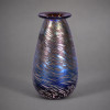 LOTTON ART GLASS — CHARLES LOTTON<br />Estados Unidos | 1988 | Vidro artístico iridescente | Assinado sob a base<br />Floreiro | 18 x 10,5 x 10,5 cm | 787 g<br /><br />Exuberante floreiro em vidro artístico iridescente, Lotton Art Glass, EUA, 1988, assinado sob a base. Corpo ovoide com gargalo em prato, decorado por traços espiralados em azul cobalto, turquesa e dourado sobre fundo rosé translúcido — técnica pulled feather com iridescência obtida por sais metálicos.<br /><br />A marca Lotton Art Glass foi fundada em 1970 por Charles Lotton, em Creta, Illinois, autodidata que revolucionou o studio glass americano. Começou no quintal; em 1973 já era referência. Charles ficou célebre por fórmulas próprias de vidro e cores únicas spot azul sobre rosa é assinatura do período 1980–1990. Em 1988, data desta peça, o estúdio já estava consolidado, com Daniel e John Lotton integrados.<br /><br />Vidro soprado à mão livre, sem molde é feito puxando fios de vidro colorido sobre a massa ainda quente, criando movimento orgânico. A iridescência vem de banhos metálicos vaporizados — estanho, titânio — que dão reflexos de azul-pavão, violeta e ouro. Assinatura gravada a diamante sob a base: Lotton 1988 — atesta autenticidade e ano.<br /><br />Lotton Art Glass integra acervos do Smithsonian Institute e Corning Museum of Glass. Charles é considerado um dos pais do American Studio Glass Movement, ao lado de Dale Chihuly e Dominick Labino. Peças assinadas, datadas, 1980s são o sweet spot do mercado: técnica madura e tamanhos expressivos.<br /><br />Vasos Lotton Art Glass, 15–20 cm, assinados, 1980s: US$ 400 a US$ 2.500 = R$ 2.200 a R$ 14.000 em leilões americanos. Com 787 g, 18 cm e iridescência intensa, esta peça situa-se entre R$ 3.500 a R$ 8.000 no Brasil, valorizada por colecionadores de studio glass.<br />
