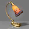 SCHNEIDER — LE VERRE FRANÇAIS<br />França | Art Déco, c. 1925–1930 | Luminária de mesa<br />Bronze dourado e cúpula em pasta de vidro assinada | 52 x 27 x 12 cm<br /><br />Excepcional luminária Art Déco da Schneider — Le Verre Français, c. 1925–1930. Haste articulada em bronze dourado com decoração vegetal rocaille, base escalonada com friso Art Déco. Cúpula campanulada em pasta de vidro dégradé, dois tons: âmbar-alaranjado ao azul-cobalto, com manchas marmorizadas típicas da manufatura. Assinada no vidro.<br /><br />Charles e Ernest Schneider fundaram em 1913 a Verrerie Schneider em Epinay-sur-Seine. Em 1918, criam a linha “Le Verre Français” — Charder para peças de luxo, Le Verre Français para produção comercial de alto nível. Rivalizaram com Daum, Muller Frères e Gallé no Art Déco francês. O vidro marmoreano em dois ou três tons, soprado e trabalhado a quente, é assinatura da casa. A produção cessa em 1938.<br /><br />A peça<br />Montagem típica Schneider: cúpula em pasta de vidro marmoreano, técnica verre jaspé, com gradação do âmbar ao azul-noturno imitando pôr do sol. Borda repicada em ondulé. Estrutura em bronze dourado col de cygne, articulada, com relevo de folhagens na haste e base pesada — desenho francês 1925, puro Art Déco. Assinatura “Le Verre Français” gravada a ácido, garantia de autenticidade.<br /><br />Schneider legítimo o Art Déco francês aceso — 1925 na sua mesa, 100 anos depois.