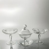 <p>Compoteira e par de pratos para doces de festa - Europa, século XX. Conjunto de três peças de cristal incolor e transparente, sendo: uma compoteira de corpo semiesférico encerrado por tampo almofadado com puxador em bola e présentoir covo. Decorada em seu todo por gravação a ácido com ramagens, pássaros e aba com faixa florida, à maneira da Cristalerie Saint Louis, 23 cm de altura total e 14,5 cm de diâmetro de boca. Par de pratos circulares e covos para doces de festa, ornados com lapidação em cata-vento e estrelada, encimando haste em balaústre facetada e base circular, 16 cm de altura total e 18 cm de diâmetro de boca. Origem: espólio de Cárbia Sabatel Bourroul e José Celestino Monteiro de Barros Bourroul.</p>
